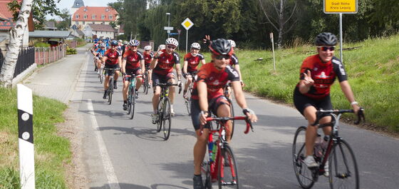 Radsportler in roter Teamkleidung fahren auf einer Dorfstraße. Einige zeigen das Victory-Zeichen.