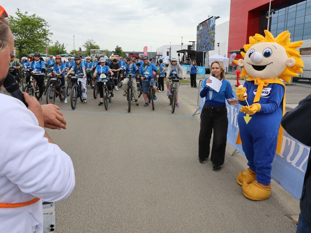 Das Maskottchen von enviaM ist mit einer Tröte in der Hand bereit, die Tour zu starten. Der moderator am linken Bildrand schaut auf die Uhr.