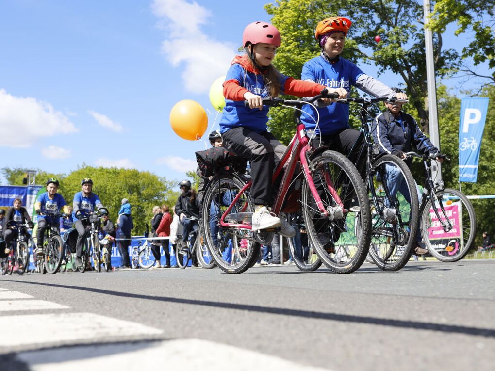 Mehrere Personen auf Fahrrädern. Die meisten tragen blaue T-Shirts. An einigen Fahrrädern sind bunte Luftballons angebunden.