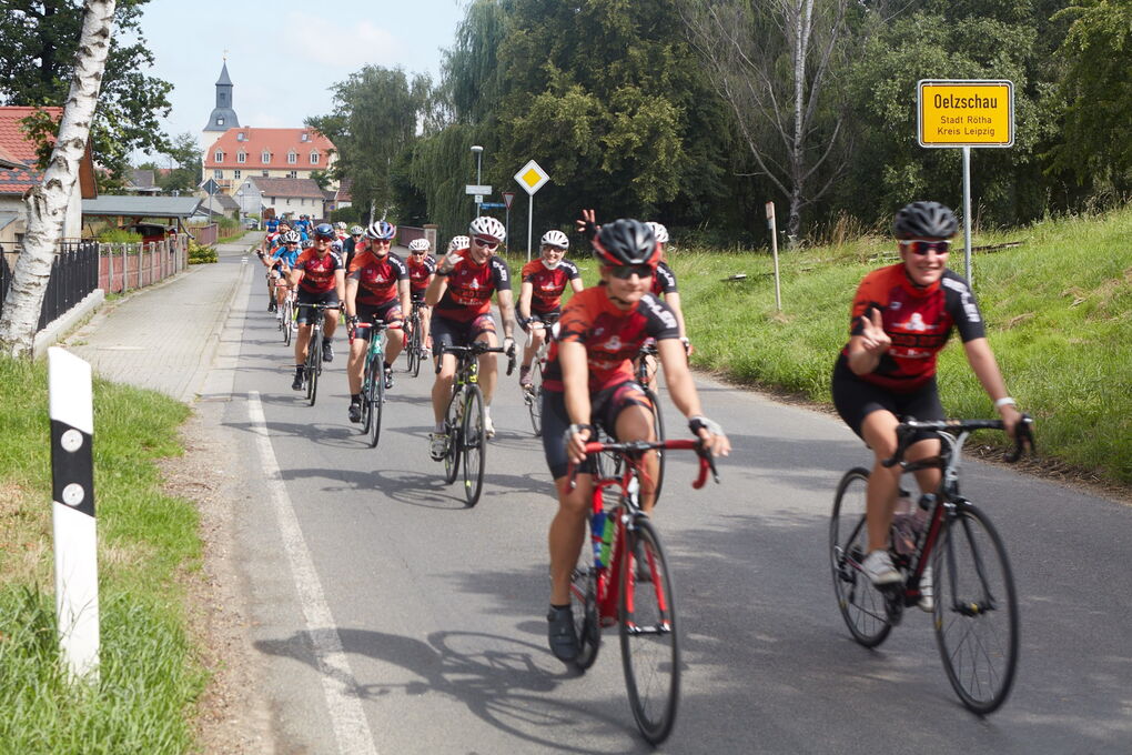 Radsportler in roter Teamkleidung fahren auf einer Dorfstraße. Einige zeigen das Victory-Zeichen.