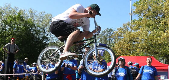 Ein BMX-Fahrer zeigt Kunststücke. Mehrere Personen in blauen Fahrradfest-Shirts schauen ihm zu.