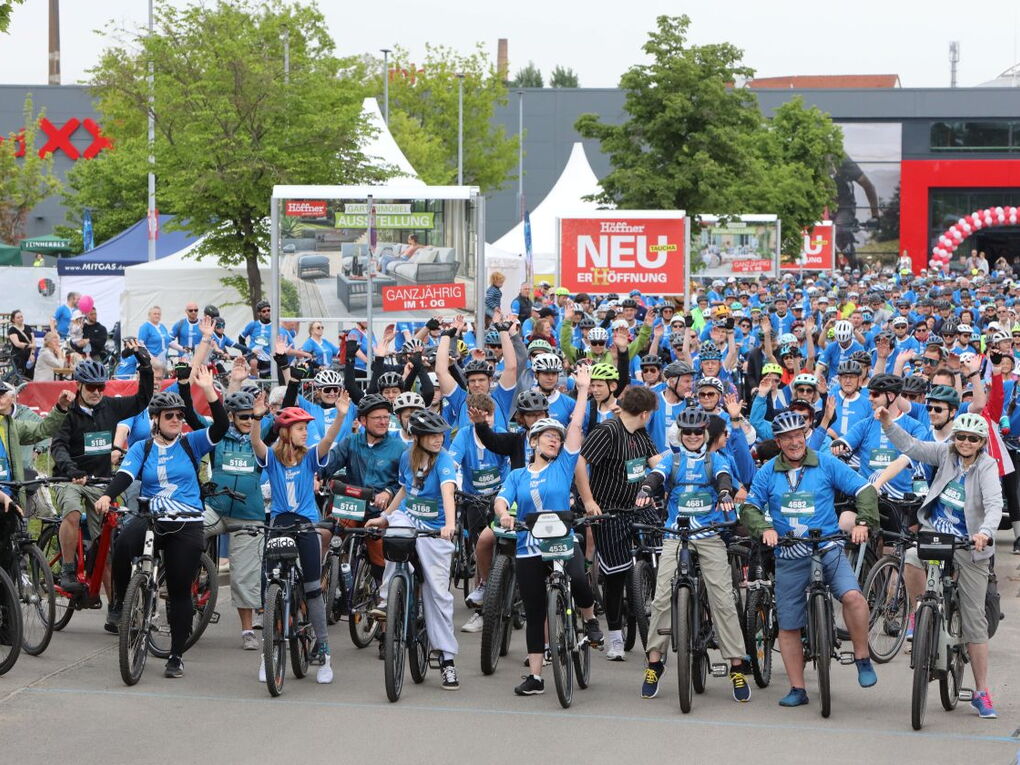 Eine große Gruppe Fahrradfahrer steht an der Startlinie, bereit zum losfahren. Sie reißen vor Begeisterung die Hände in die Luft.