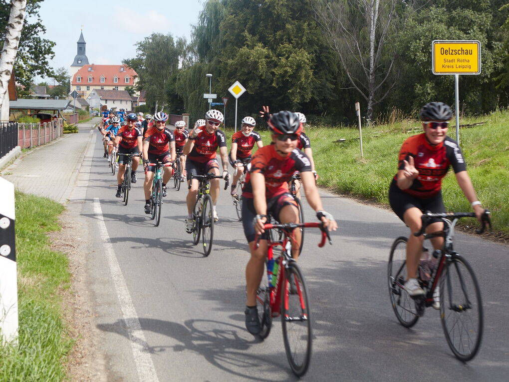 Radsportler in roter Teamkleidung fahren auf einer Dorfstraße. Einige zeigen das Victory-Zeichen.