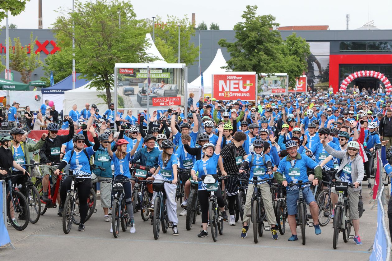 Eine große Gruppe Fahrradfahrer steht an der Startlinie, bereit zum losfahren. Sie reißen vor Begeisterung die Hände in die Luft.