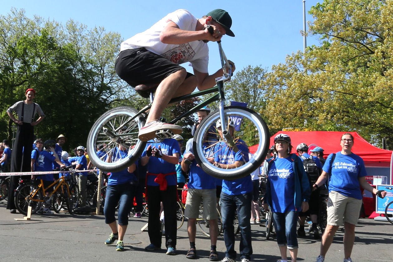 Ein BMX-Fahrer zeigt Kunststücke, mehrere Menschen in blauen Fahrradfest-T-Shirts schauen zu.
